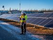 Indigenous solar technician with tablet beside a ground-mounted solar array in a northern Canadian community at golden hour, with a small communications tower, boreal forest, and a rooftop satellite dish in the background.