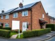UK suburban brick semi-detached house with rooftop solar panels and a blank real estate sale sign in the front garden, shot at eye level on a bright overcast day, with neighboring houses and hedges softly blurred in the background.
