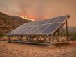 Fire-resilient solar panels mounted on a raised frame in a dry hillside landscape with smoky wildfire haze in the distance at sunset.